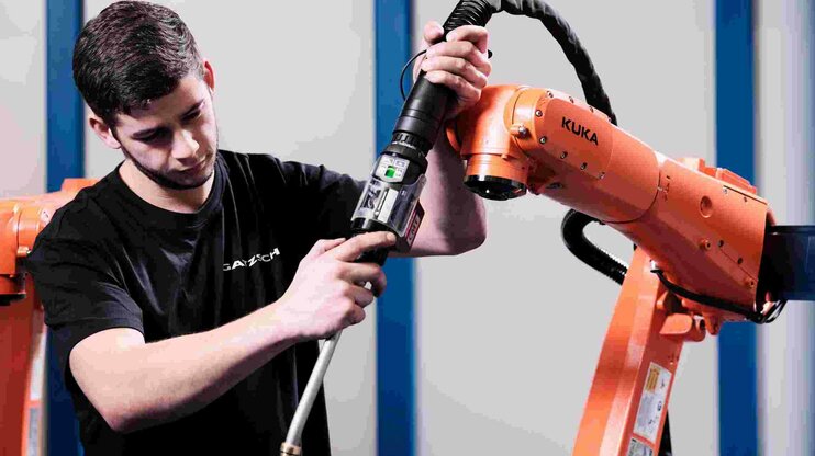 Ein junger Industriemechaniker arbeitet an einem orangefarbenen KUKA-Roboterarm. Der Techniker hält ein Werkzeug in der Hand und führt Wartungs- oder Installationsarbeiten durch. Im Hintergrund sind weitere Roboterarme zu sehen, die auf den industriellen Einsatz in der Produktion hinweisen. Die Szene steht für die technische Expertise und den Einsatz modernster Robotik in der Schweißtechnik bei Gatzsch. Sie symbolisiert die Arbeit von Fachkräften, die dafür sorgen, dass automatisierte Fertigungssysteme präzise und zuverlässig funktionieren.