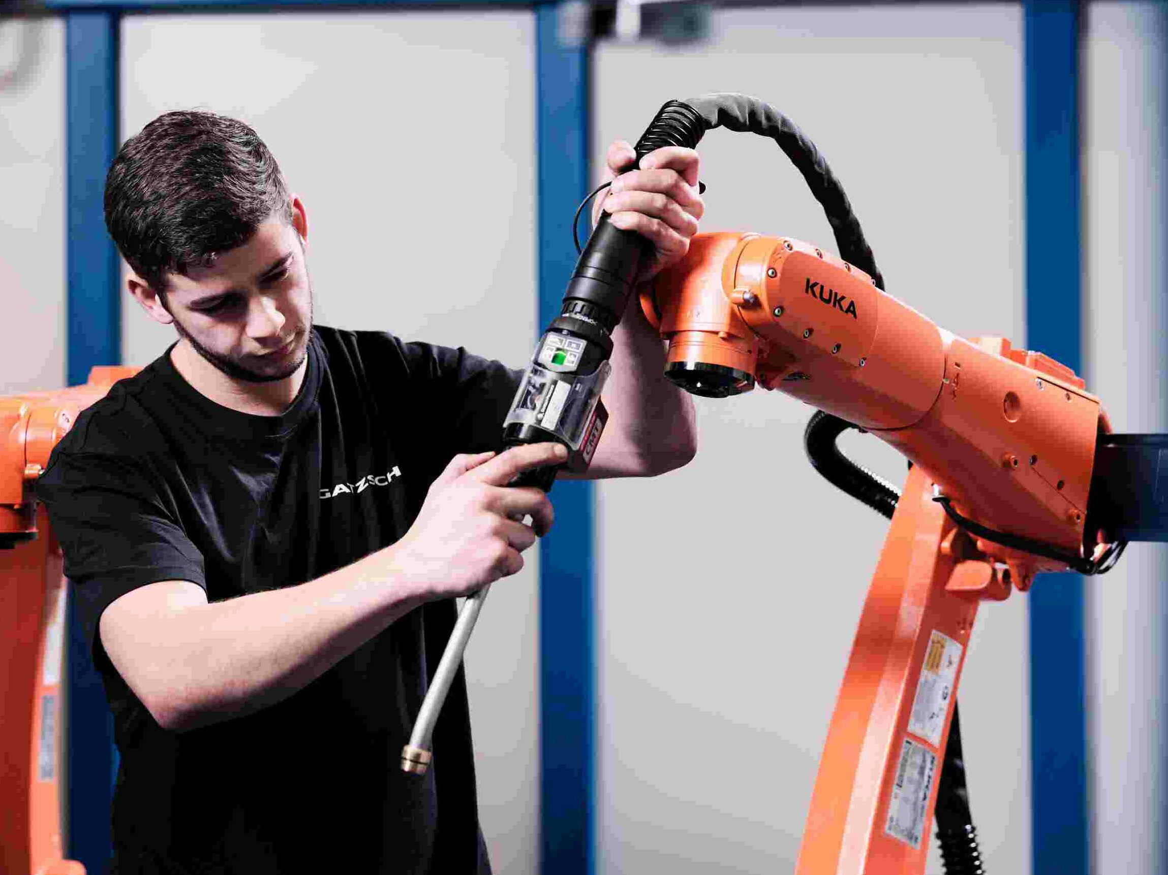 Ein junger Industriemechaniker arbeitet an einem orangefarbenen KUKA-Roboterarm. Der Techniker hält ein Werkzeug in der Hand und führt Wartungs- oder Installationsarbeiten durch. Im Hintergrund sind weitere Roboterarme zu sehen, die auf den industriellen Einsatz in der Produktion hinweisen. Die Szene steht für die technische Expertise und den Einsatz modernster Robotik in der Schweißtechnik bei Gatzsch. Sie symbolisiert die Arbeit von Fachkräften, die dafür sorgen, dass automatisierte Fertigungssysteme präzise und zuverlässig funktionieren.