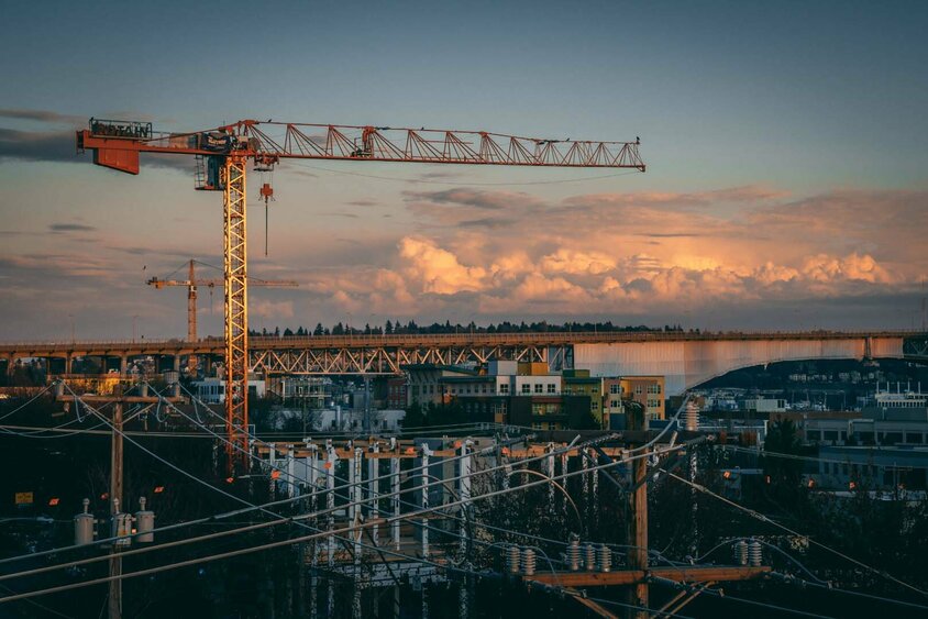  Eine Baustelle im Sonnenuntergang, auf der ein großer Baukran über einem Stadtgebiet schwebt. Im Hintergrund sind Gebäude, eine Brücke und Wolkenformationen zu sehen. Die Szene steht symbolisch für die Bauindustrie, eine Branche, die von der Gatzsch Schweißtechnik mit industriellen Fügetechniken beliefert wird. Der Kran repräsentiert die Bedeutung robuster und präziser Schweißverbindungen in der Bau- und Konstruktionsindustrie.