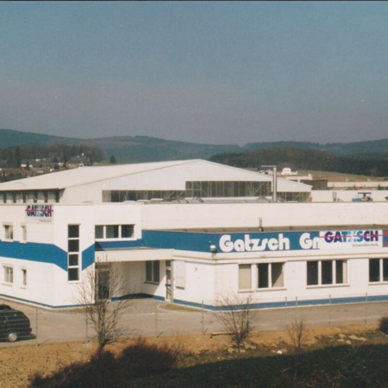 Historische Außenansicht des Gatzsch Firmengebäudes im Jahr 1979. Das Gebäude ist in einem weißen Farbton mit blauen Streifen gehalten und trägt das Gatzsch-Logo auf der Fassade. Es handelt sich um ein zweistöckiges Büro- und Produktionsgebäude mit angeschlossener Halle. Im Vordergrund sind geparkte Autos und eine umzäunte Einfahrt zu sehen. Im Hintergrund erstrecken sich Hügel und eine ländliche Umgebung. Das Bild zeigt die frühen Jahre des Unternehmens und seine Entwicklung im Bereich der Schweißtechnik.