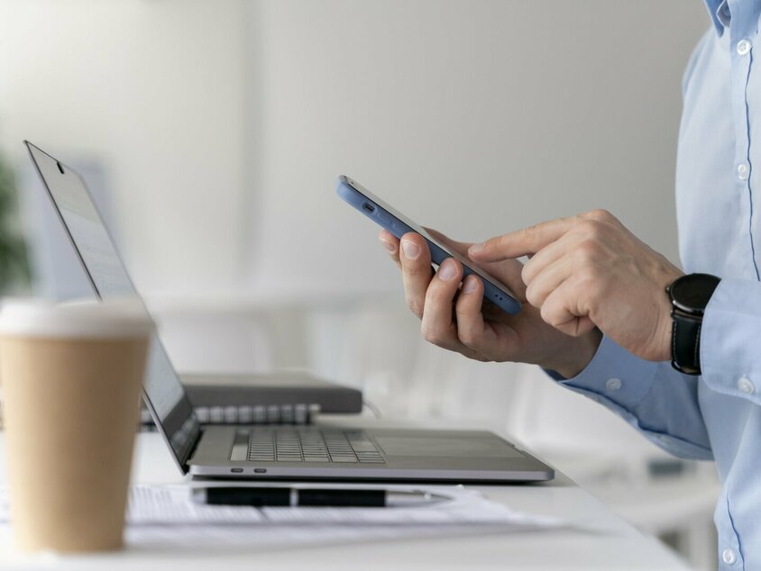 Ein Mitarbeiter sitzt an einem Schreibtisch und bedient ein Smartphone, während ein Laptop vor ihm geöffnet ist. Auf dem Tisch liegt ein Kaffeebecher und Notizmaterialien. Diese Szene symbolisiert den After-Sales-Service bei Gatzsch Schweißtechnik, der moderne Kommunikationsmittel nutzt, um den Kunden schnellen und zuverlässigen Support zu bieten.