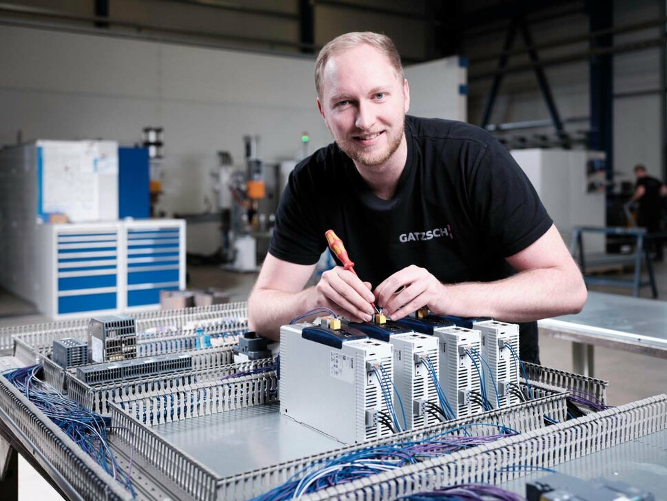 An electrician from Gatzsch Schweißtechnik is working on a control cabinet in a production hall, wiring electronic components. The employee is wearing a black T-shirt with the company logo and smiling at the camera. Tools and technical equipment can be seen in the background. The image symbolizes the precise work of the electricians in wiring and installing welding systems.