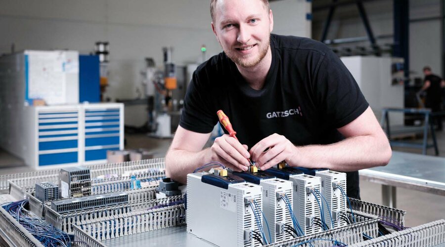 Ein Elektriker der Firma Gatzsch Schweißtechnik arbeitet in einer Produktionshalle an einem Schaltschrank und verkabelt elektronische Komponenten. Der Mitarbeiter trägt ein schwarzes T-Shirt mit dem Firmenlogo und lächelt in die Kamera. Im Hintergrund sind Werkzeuge und technische Ausrüstungen zu erkennen. Das Bild symbolisiert die präzise Arbeit der Elektriker bei der Verkabelung und Installation von Schweißanlagen.