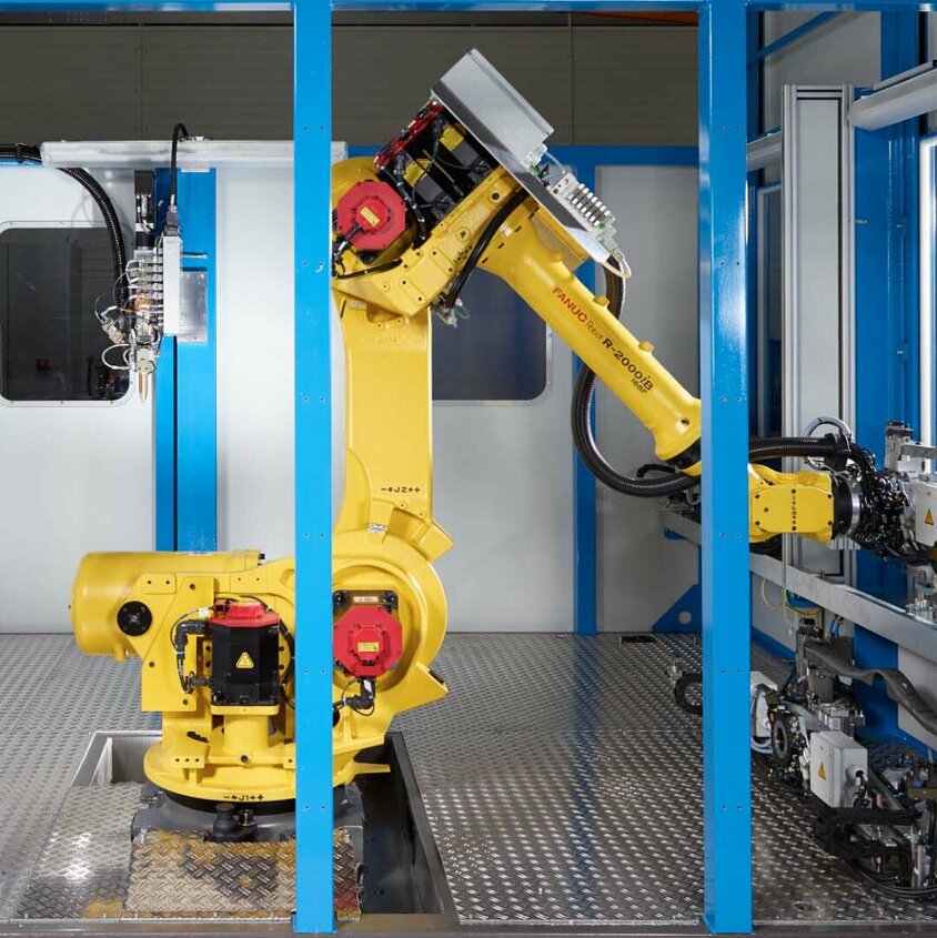 Automatisierte Klebetechnik: Präzision und Effizienz in der Fertigung bei Gatzsch Schweißtechnik Eine industrielle Klebeanlage mit einem gelben FANUC-Roboterarm in einem abgeschirmten Arbeitsbereich. Der Roboter ist in der Mitte der Anlage positioniert, bereit für automatisierte Aufgaben. Umgeben von blauen Rahmenstrukturen, zeigt die Szene eine hochmoderne Fertigungsumgebung mit mechanischen Komponenten und Steuergeräten. Diese Klebeanlage steht für Präzision, Effizienz und Automatisierung in der Produktion, wie sie bei Gatzsch Schweißtechnik eingesetzt wird.