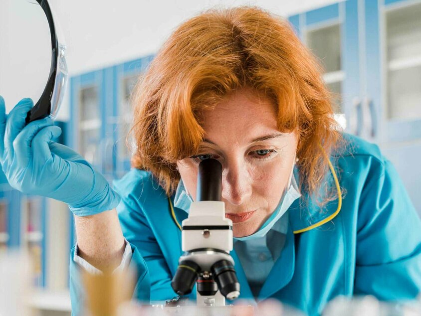 Eine Frau mit rotem Haar und blauer Arbeitskleidung blickt durch ein Mikroskop in einem Labor. Sie hält eine Schutzbrille in einer Hand und trägt blaue Einweghandschuhe. Im Vordergrund sind unscharfe Laborproben zu erkennen. Der Hintergrund zeigt Schränke und Labormaterialien, was auf eine wissenschaftliche Umgebung hinweist, möglicherweise ein Schweißlabor im Technologiecenter der Gatzsch Schweißtechnik.