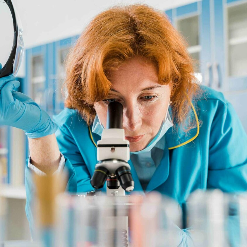 Eine Frau mit rotem Haar und blauer Arbeitskleidung blickt durch ein Mikroskop in einem Labor. Sie hält eine Schutzbrille in einer Hand und trägt blaue Einweghandschuhe. Im Vordergrund sind unscharfe Laborproben zu erkennen. Der Hintergrund zeigt Schränke und Labormaterialien, was auf eine wissenschaftliche Umgebung hinweist, möglicherweise ein Schweißlabor im Technologiecenter der Gatzsch Schweißtechnik.