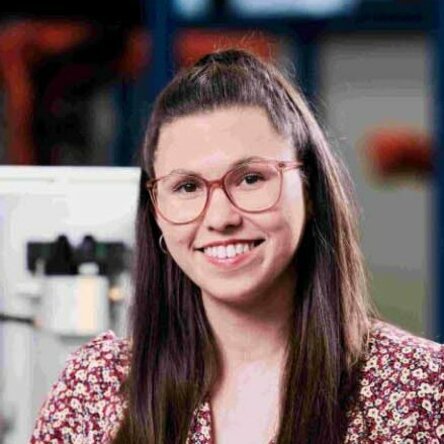 Ms. Funke, Head of Accounting at Gatzsch Schweißtechnik, stands in an industrial setting next to a technical device. She is wearing a red patterned blouse and jeans. She leans against the device with one hand and smiles warmly at the camera. Machines and equipment can be seen in the background.