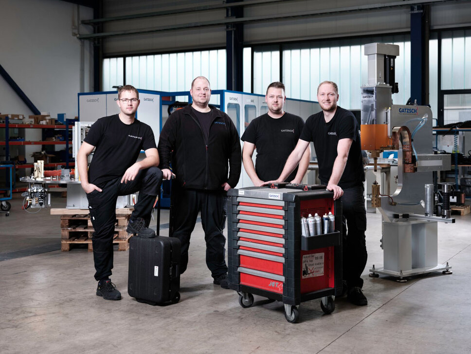 Four employees from Gatzsch Schweißtechnik stand in a workshop in front of welding systems. They wear black work clothes with the Gatzsch logo and appear ready for technical service. One employee is leaning on a tool cart, while another is standing on a case. In the background, welding systems and workbenches can be seen, indicating a well-equipped production environment.