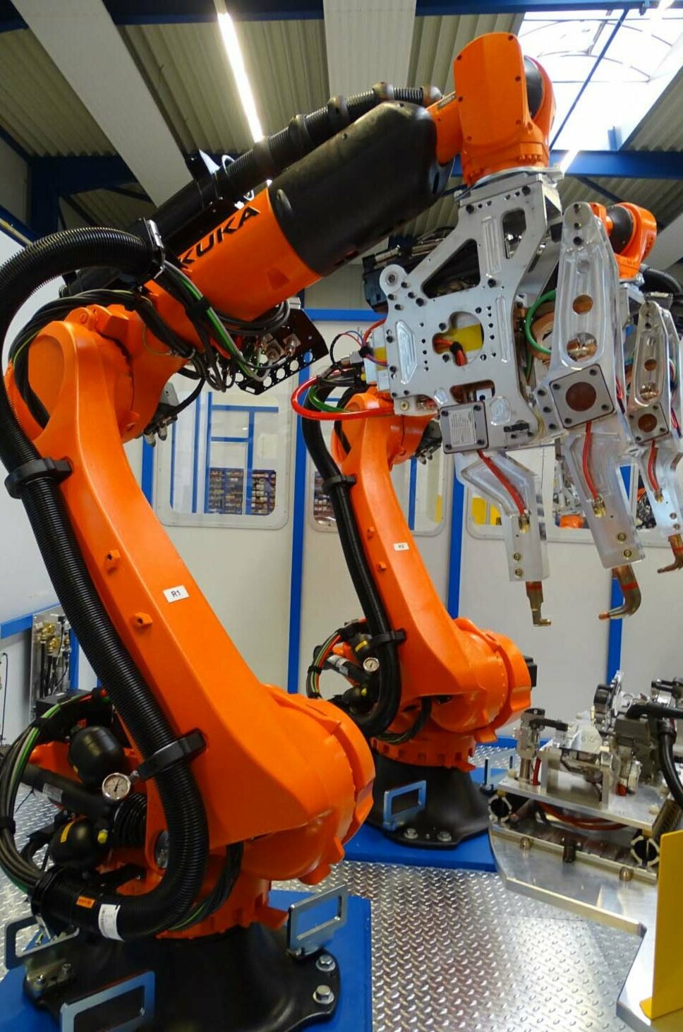 Das Bild zeigt einen orangefarbenen Industrieroboterarm in einer Fabrikumgebung, der für automatisiertes Punktschweißen eingesetzt wird. Der Roboter greift mit seinen mechanischen Greifarmen ein Bauteil, um präzise Schweißpunkte anzubringen. Die Anlage ist Teil einer modernen Produktionslinie, die Schweißprozesse effizient automatisiert.