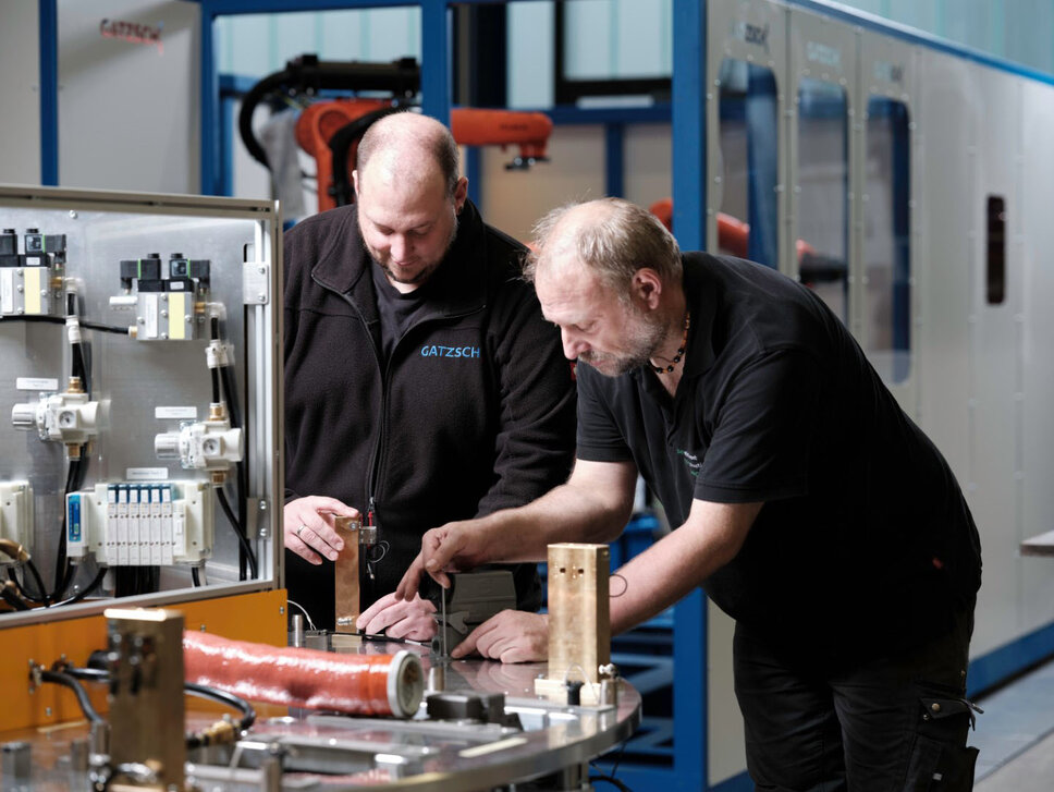 Cooperation and expertise in system assembly: commissioning complex welding systems at Gatzsch Schweißtechnik wo employees of Gatzsch Schweißtechnik work together on a welding system during the assembly process. They are reviewing and discussing the components of the welding system, with one employee pointing to a specific element. In the background, a modern production hall with more machines can be seen. The image conveys the collaboration and expertise required for the commissioning and maintenance of complex welding systems.