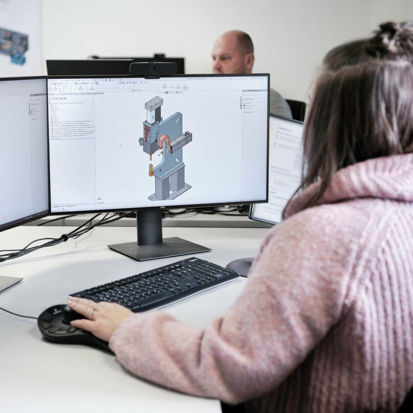 Eine Mitarbeiterin der Firma Gatzsch Schweißtechnik arbeitet an einem Computer mit zwei Monitoren, auf denen 3D-Modelle von Schweißanlagen dargestellt sind. Sie trägt einen rosa Pullover und konzentriert sich auf die Konstruktionssoftware, während sie mit einer 3D-Maus arbeitet. Im Hintergrund ist ein weiterer Kollege ebenfalls mit der Konstruktion beschäftigt. Das Bild vermittelt einen Einblick in die präzise Entwicklungsarbeit der Ingenieure und Konstrukteure bei Gatzsch.