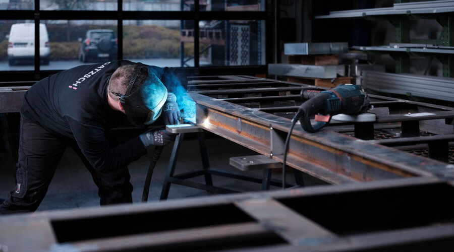 Ein Mitarbeiter der Firma Gatzsch Schweißtechnik ist in einer Werkstatt beim Schweißen zu sehen. Er trägt eine Schweißmaske und arbeitet konzentriert an einem großen Metallrahmen, während blauer Lichtschein vom Schweißvorgang zu sehen ist. Auf dem Tisch neben ihm liegt ein Winkelschleifer. Im Hintergrund sind Fenster zu sehen, die den Blick auf Fahrzeuge und eine Außenansicht der Umgebung freigeben. Das Bild zeigt die präzise und handwerklich anspruchsvolle Metallbauarbeit bei Gatzsch.