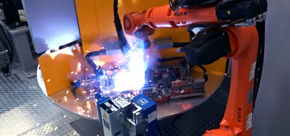 Ein Roboterarm führt einen Lichtbogenschweißvorgang durch, wobei helle Funken sprühen und Metallstücke in einer automatisierten Fertigungsumgebung miteinander verschweißt werden.