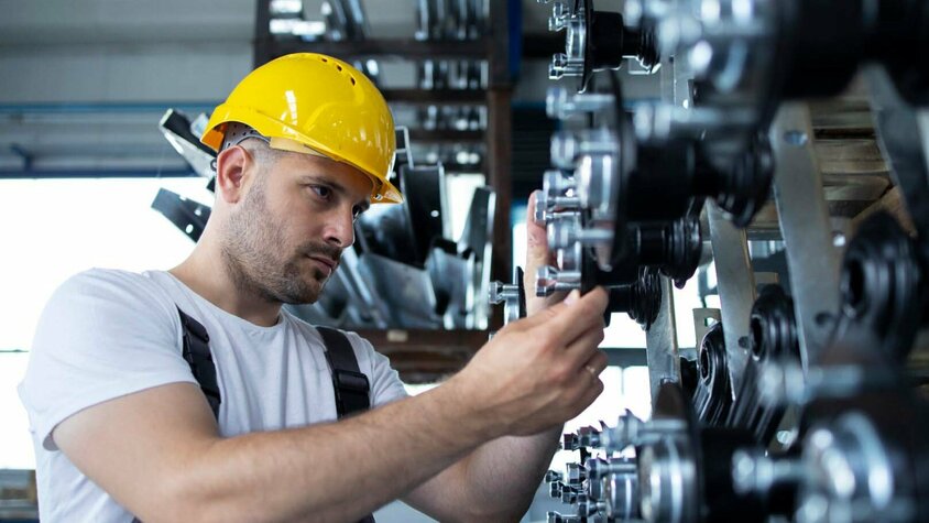 Ein Arbeiter mit gelbem Schutzhelm überprüft Maschinenteile in einer industriellen Fertigungsumgebung. Er arbeitet präzise an einer Reihe von Metallkomponenten. Das Bild symbolisiert die Maschinenbauindustrie, in die Gatzsch Schweißtechnik Anlagen zum Nieten und Clinchen liefert.