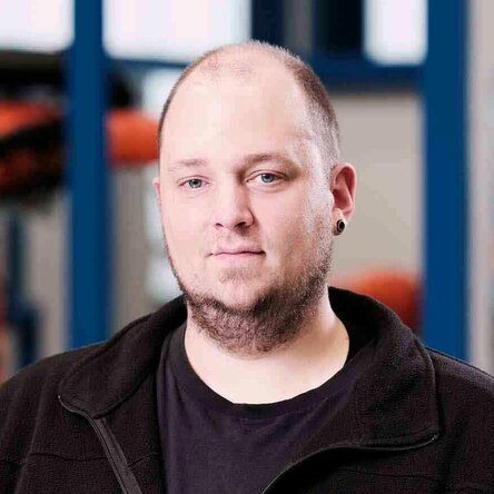 Mr. Matrose, Head of Production at Gatzsch Schweißtechnik, stands in a technical work environment. He is wearing a black fleece jacket with the Gatzsch logo. Industrial machines and equipment are visible in the background. Mr. Matrose looks calmly into the camera while leaning on a technical device with one hand.