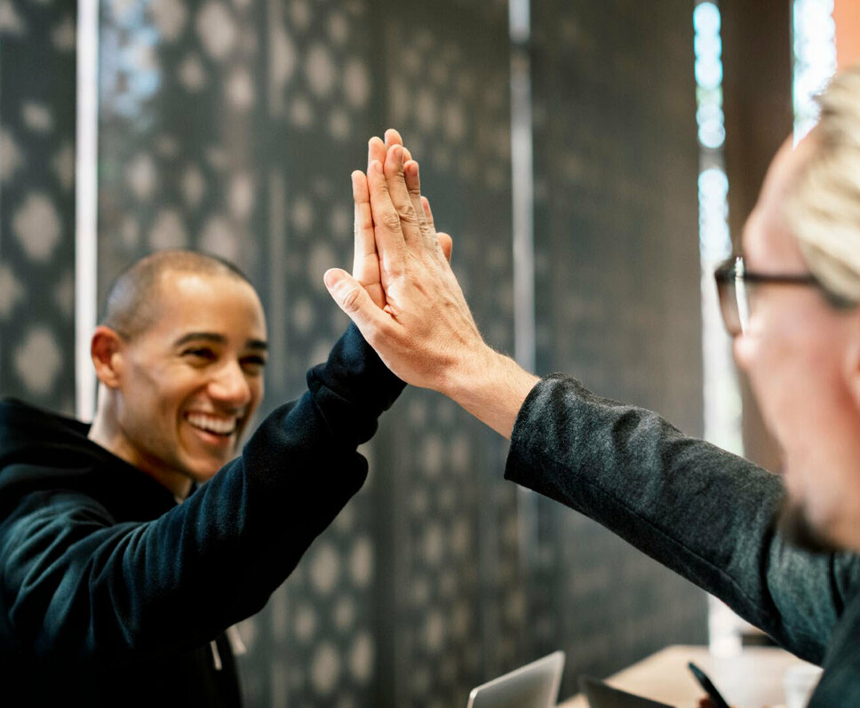 Zwei Kollegen geben sich in einem Büro High-Five, beide lächeln und wirken zufrieden. Die Szene symbolisiert Erfolg, Zusammenarbeit und eine positive Arbeitsatmosphäre. Im Hintergrund sind verschwommene Büroelemente zu sehen, die auf eine professionelle Umgebung hinweisen. Das Bild steht für die Vorteile und den Teamgeist, die Mitarbeiter bei Gatzsch Schweißtechnik in Attendorn erleben, und verdeutlicht die Bedeutung von gegenseitiger Unterstützung und Freude an der Arbeit.