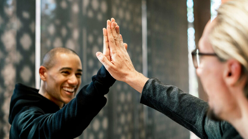 Zwei Kollegen geben sich in einem Büro High-Five, beide lächeln und wirken zufrieden. Die Szene symbolisiert Erfolg, Zusammenarbeit und eine positive Arbeitsatmosphäre. Im Hintergrund sind verschwommene Büroelemente zu sehen, die auf eine professionelle Umgebung hinweisen. Das Bild steht für die Vorteile und den Teamgeist, die Mitarbeiter bei Gatzsch Schweißtechnik in Attendorn erleben, und verdeutlicht die Bedeutung von gegenseitiger Unterstützung und Freude an der Arbeit.