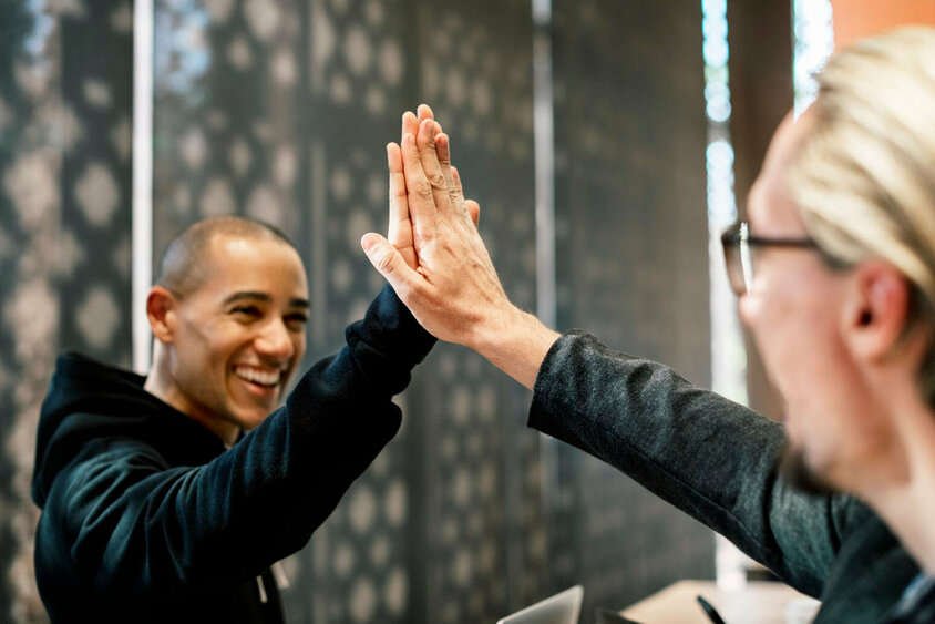 Zwei Kollegen geben sich in einem Büro High-Five, beide lächeln und wirken zufrieden. Die Szene symbolisiert Erfolg, Zusammenarbeit und eine positive Arbeitsatmosphäre. Im Hintergrund sind verschwommene Büroelemente zu sehen, die auf eine professionelle Umgebung hinweisen. Das Bild steht für die Vorteile und den Teamgeist, die Mitarbeiter bei Gatzsch Schweißtechnik in Attendorn erleben, und verdeutlicht die Bedeutung von gegenseitiger Unterstützung und Freude an der Arbeit.