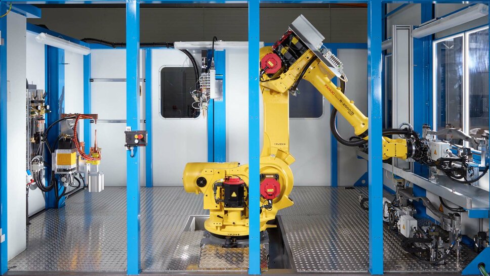 Eine industrielle Klebeanlage mit einem gelben FANUC-Roboterarm in einem abgeschirmten Arbeitsbereich. Der Roboter ist in der Mitte der Anlage positioniert, bereit für automatisierte Aufgaben. Umgeben von blauen Rahmenstrukturen, zeigt die Szene eine hochmoderne Fertigungsumgebung mit mechanischen Komponenten und Steuergeräten. Diese Klebeanlage steht für Präzision, Effizienz und Automatisierung in der Produktion, wie sie bei Gatzsch Schweißtechnik eingesetzt wird.