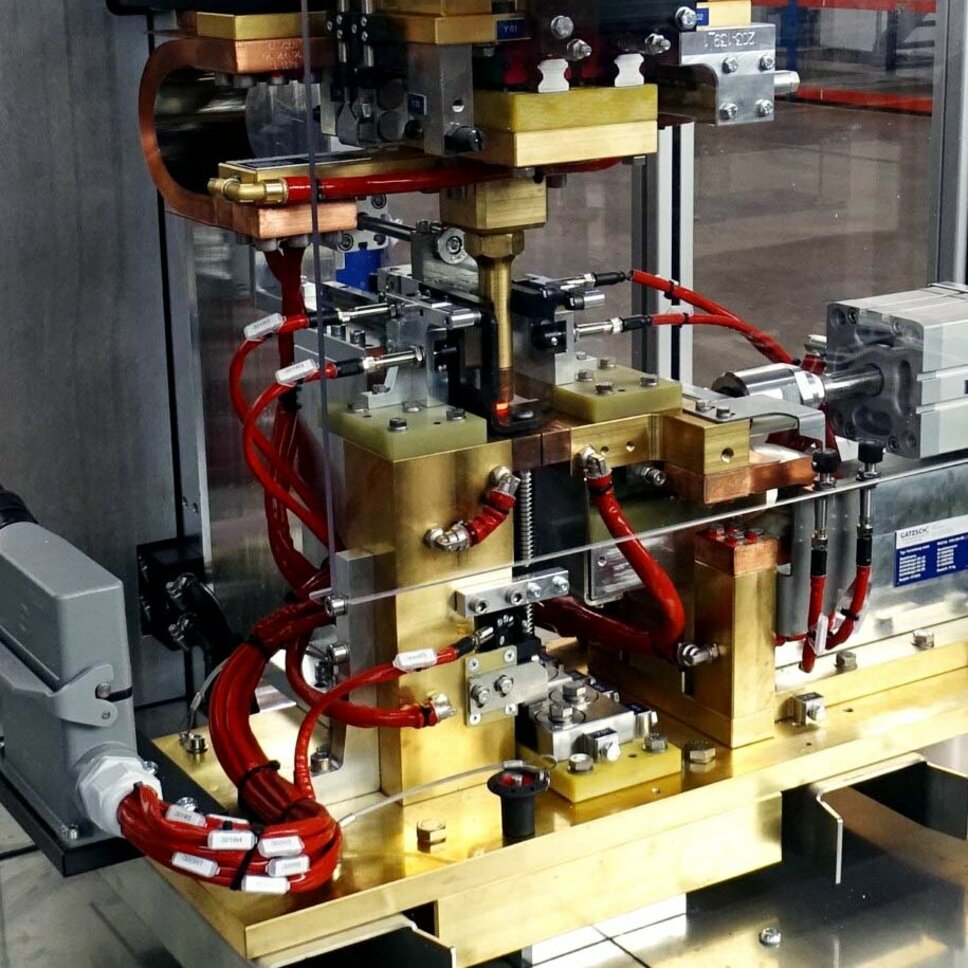 Industrielle Warmnietmaschine in einer Produktionsumgebung. Die Maschine besteht aus komplexen Komponenten, darunter elektrische Leitungen, hydraulische Zylinder und Metallwerkzeuge, die auf ein Werkstück ausgerichtet sind. Das Gerät trägt das Logo von Gatzsch und zeigt eine robuste Bauweise, die auf präzises Warmnieten spezialisiert ist, um zuverlässige und sichere Verbindungen in der Fertigung herzustellen.