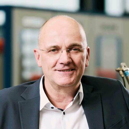 Mr. Gatzsch, Managing Director of Gatzsch Schweißtechnik, stands in an industrial environment next to a welding machine with the Gatzsch logo. He is wearing a dark gray suit with a white shirt and is smiling at the camera. He leans casually against the machine with one hand. Production equipment can be seen in the background.