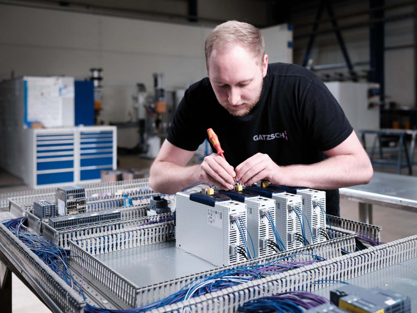 Ein Elektriker der Firma Gatzsch Schweißtechnik arbeitet konzentriert an der Verdrahtung einer Steuereinheit in der Anlagenmontage. Er nutzt einen Schraubendreher, um Verbindungen in einem Schaltschrank zu fixieren. Die Umgebung deutet auf eine Produktionshalle hin, in der verschiedene Baugruppen und Schaltschränke montiert werden. Das Bild zeigt das präzise und sorgfältige Arbeiten, das für den Aufbau und die Verkabelung von Schweißanlagen unerlässlich ist.